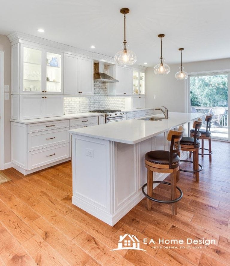 A view of a renovated kitchen with a focus on the appliances. A large, white refrigerator is visible on the far left, while a white microwave and stove stand out in the center and to the right. The countertops are a light, solid color, complementing the creamy white cabinets.