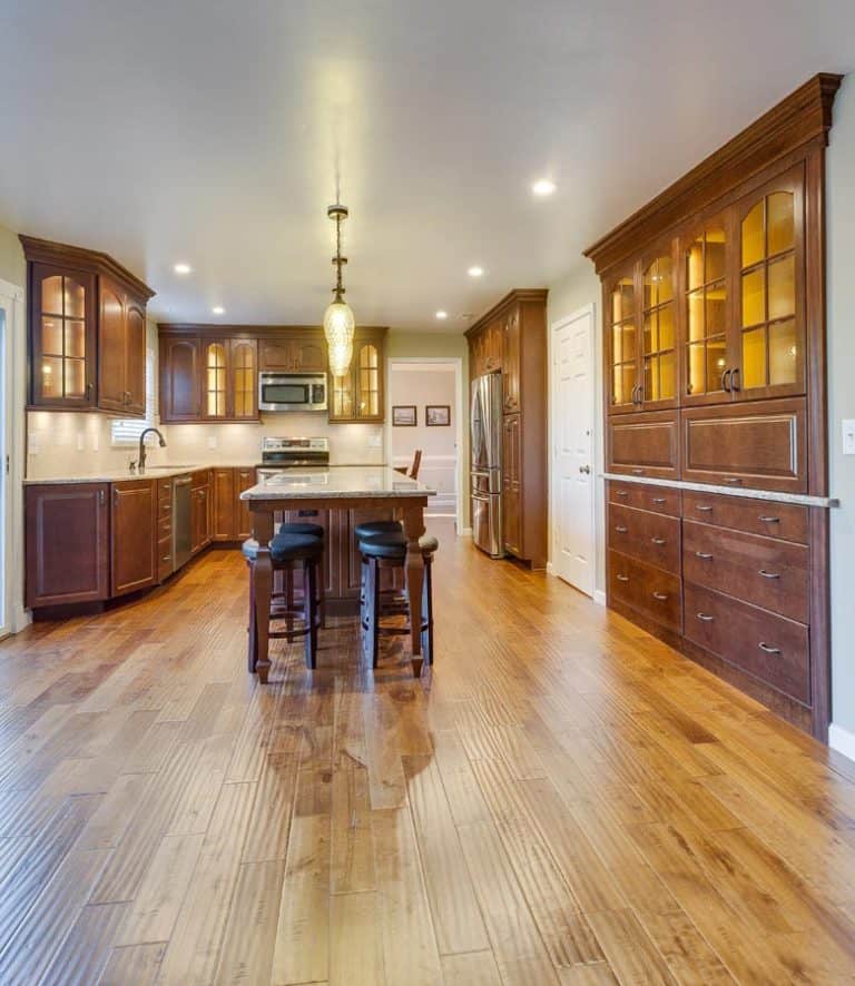 A shot capturing the warm tones of a kitchen's interior design. The glossy, reddish-brown hardwood floor complements the dark cherry wood of the cabinets and island. The light countertop of the island and the stainless steel refrigerator provide a modern contrast to the traditional wood.