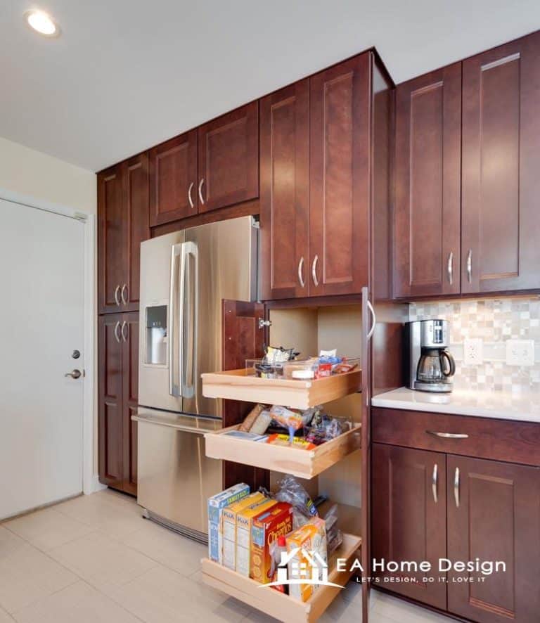 A photograph highlighting the appliances and cabinetry in a residential kitchen. The built-in stainless steel refrigerator is the focal point on the right, surrounded by floor-to-ceiling dark wood cabinets. The image also shows a wine rack and upper cabinets above the island.