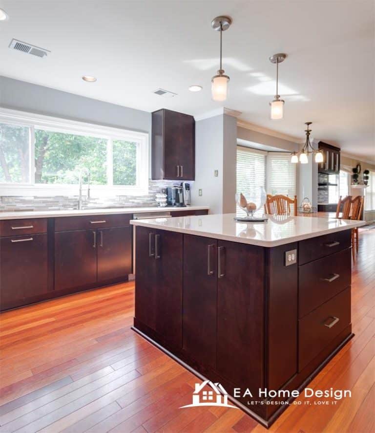 An interior photograph of a renovated kitchen space. The image is taken from a low viewpoint, looking across the large kitchen island. The island features multiple cabinet doors and a white or light marble countertop. Behind the island, a massive stainless steel fridge is integrated into the cabinetry.