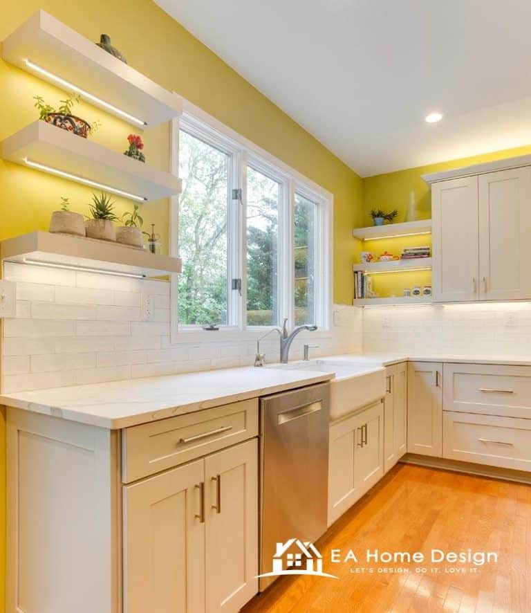 A view of a renovated kitchen with a focus on the appliances. A large, white refrigerator is visible on the far left, while a white microwave and stove stand out in the center and to the right. The countertops are a light, solid color, complementing the creamy white cabinets.