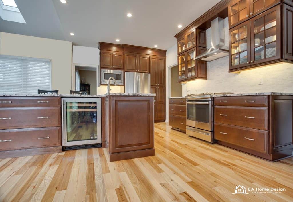 An interior image capturing the natural light in a kitchen. A large bay window on the left with blinds allows light to flood the room, illuminating the wood floors and granite countertops. The space is a blend of a kitchen and a dining area, creating a functional, open environment.