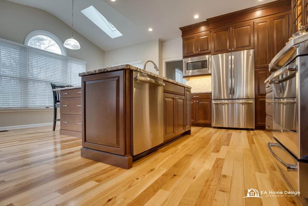 A photograph from the perspective of someone entering the kitchen. The foreground shows a white door and part of a light-colored wall, drawing the eye into the warm space. The kitchen features wood cabinetry, a breakfast bar, and a dining table at the far end.