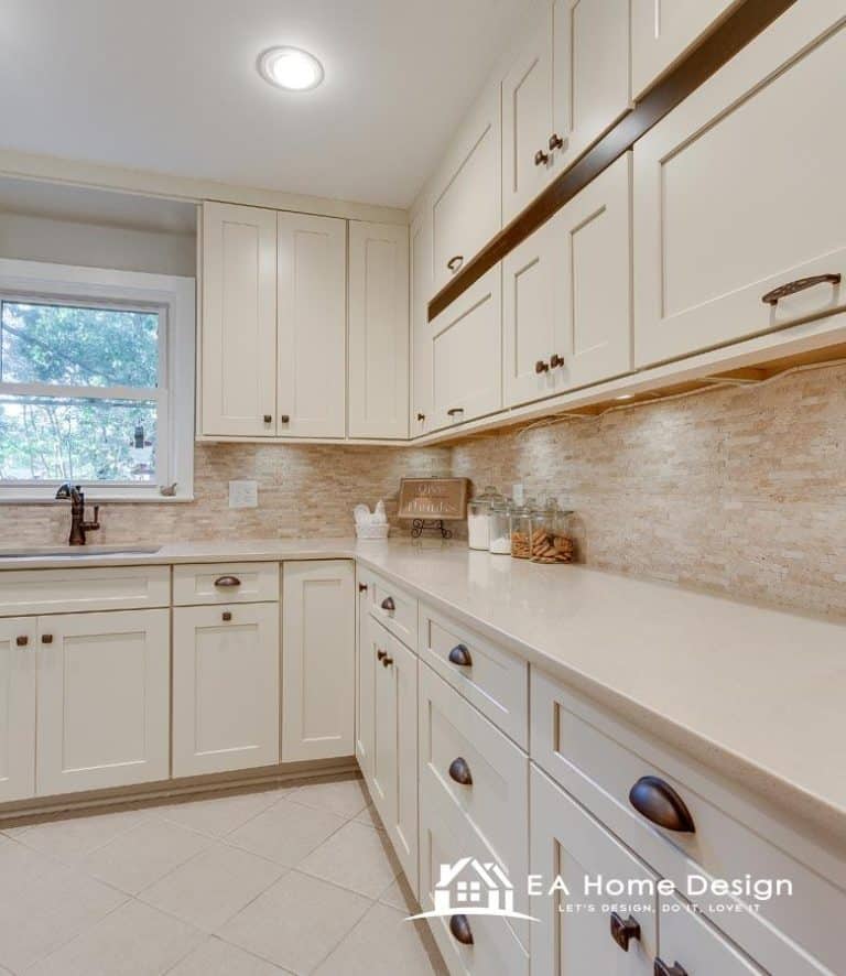An image capturing the minimalist design of a bright kitchen. The walls are lined with off-white shaker-style cabinets and a textured stone or brick backsplash. The floor is tiled with light-colored, square tiles, creating a cohesive, clean look.