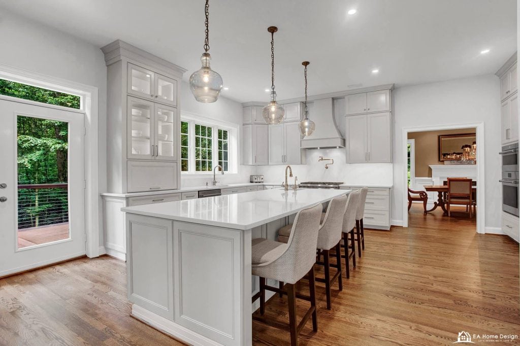 A professional photograph of a spacious, modern kitchen with a large white quartz island. The kitchen features high-end stainless steel appliances, including a double-door refrigerator and a built-in oven and microwave, set into tall white cabinets.