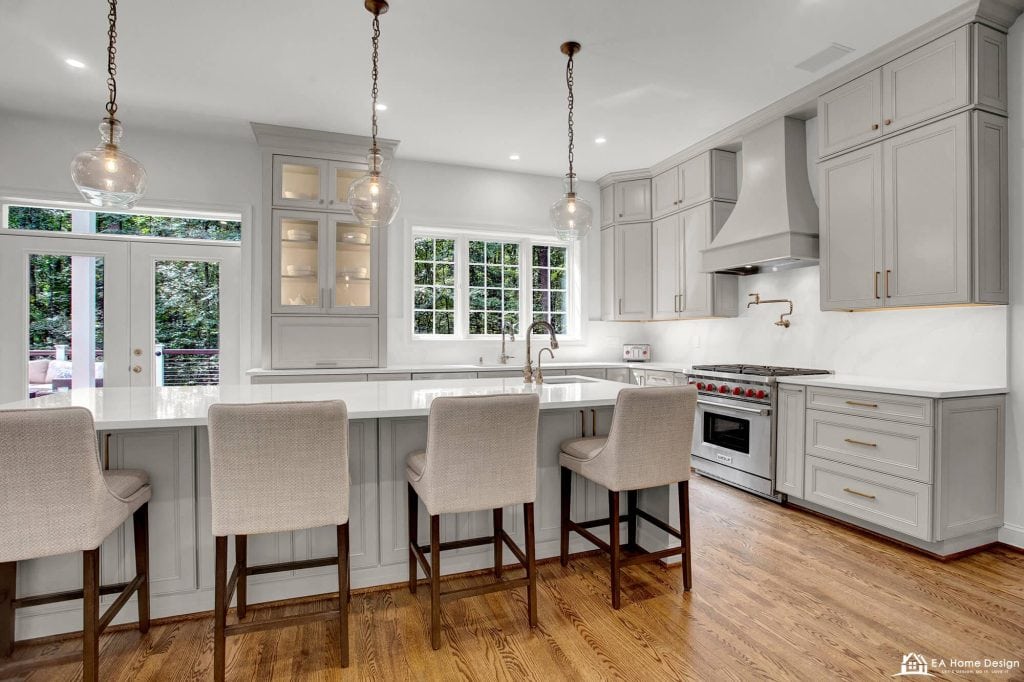 A professional photograph of a spacious, modern kitchen with a large white quartz island. The kitchen features high-end stainless steel appliances, including a double-door refrigerator and a built-in oven and microwave, set into tall white cabinets.