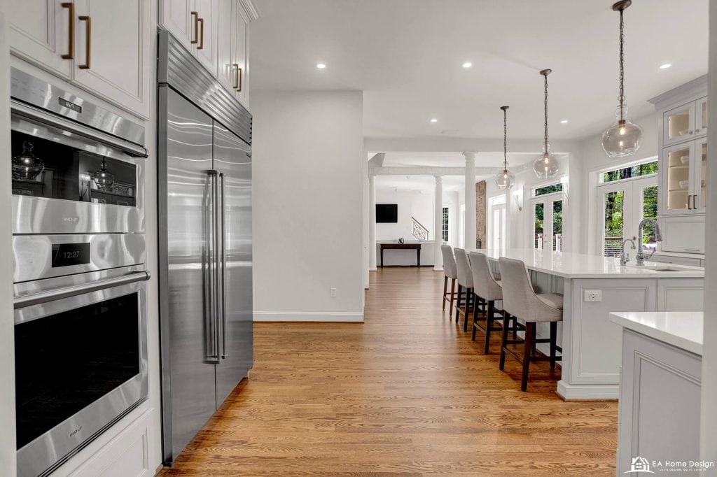 A professional photograph of a spacious, modern kitchen with a large white quartz island. The kitchen features high-end stainless steel appliances, including a double-door refrigerator and a built-in oven and microwave, set into tall white cabinets.
