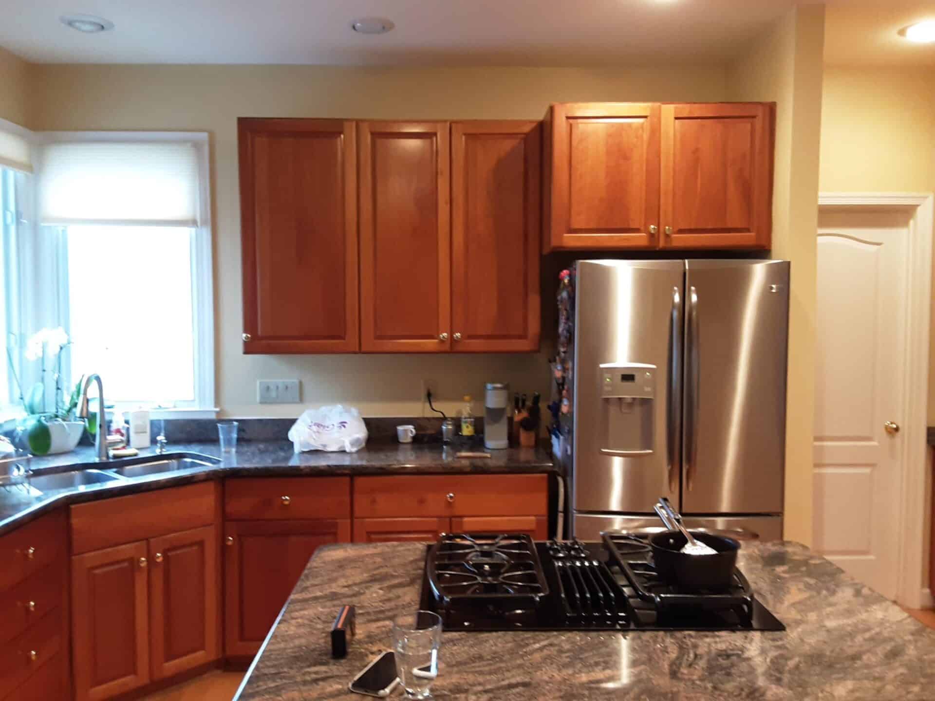 A Northern Virginia kitchen before its remodel by EA Home Design: cherry cabinets, a dark granite counter, a stainless refrigerator standing well proud of the counter beside the door, and a peninsula with a cooktop crowding the walkway