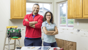 Two people in a kitchen under renovation with tools and cabinets visible.