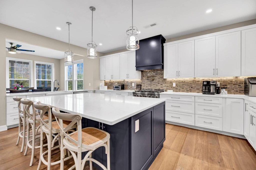 A wide view of a modern kitchen with sleek appliances, a large dining table, a sink under a window, and recessed ceiling lights.