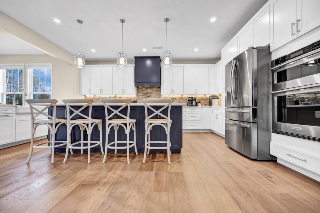A modern kitchen from the dining area, showcasing a large fridge, a gas stove, a farmhouse sink, and elegant light fixtures.