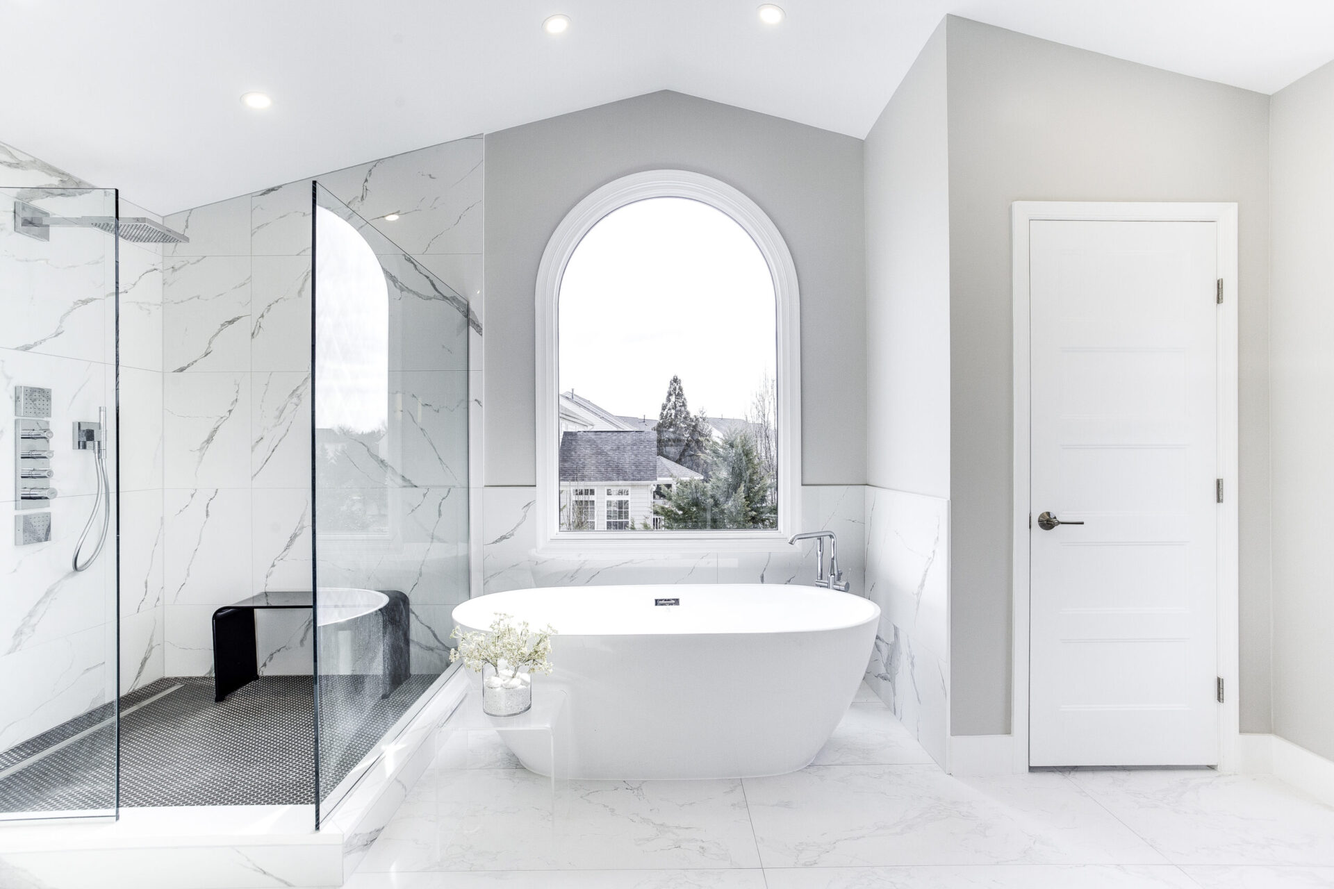White marble bathroom with a tall arched window over a freestanding tub, a marble walk in shower and veined marble walls