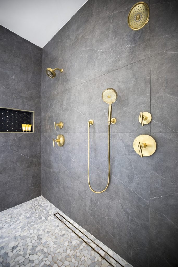 After: inside the shower, dark gray large format tile, gold rain head, hand shower and body sprays, recessed niche, mosaic floor with linear drain