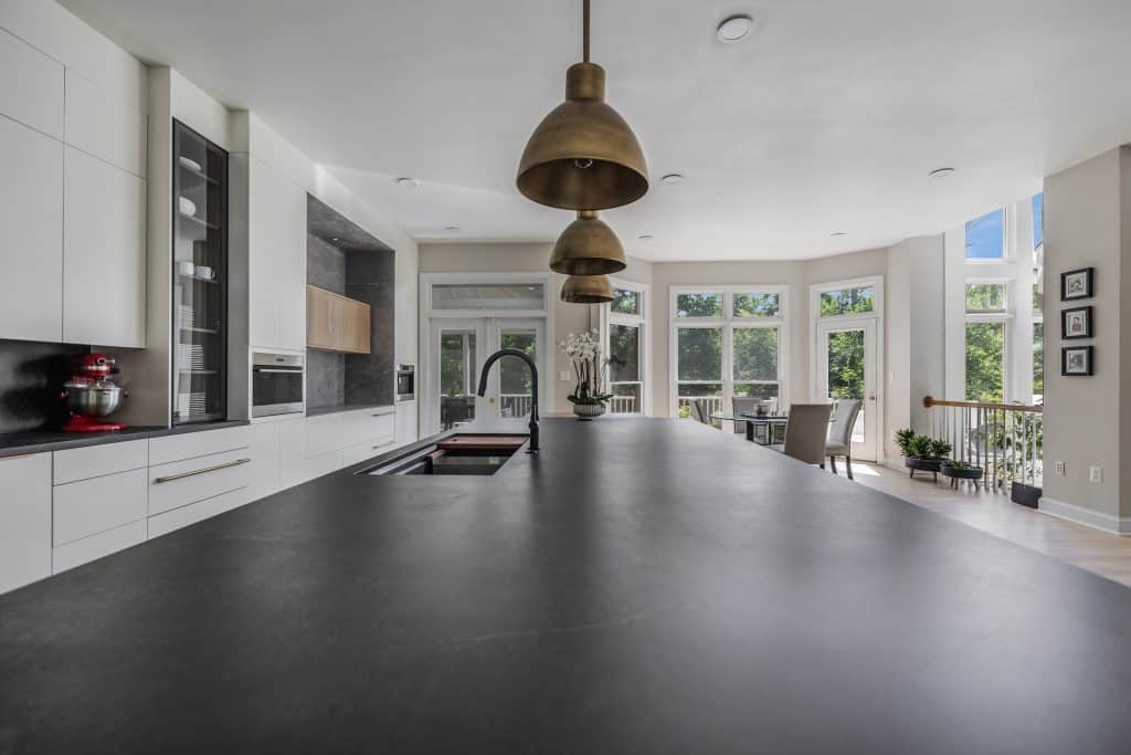 This contemporary kitchen combines white cabinetry with black countertops and sleek modern finishes. A large bay window fills the space with natural light, highlighting the central island topped with stylish pendant lighting. The kitchen seamlessly connects to an open dining area, creating an airy, functional, and inviting space ideal for modern living