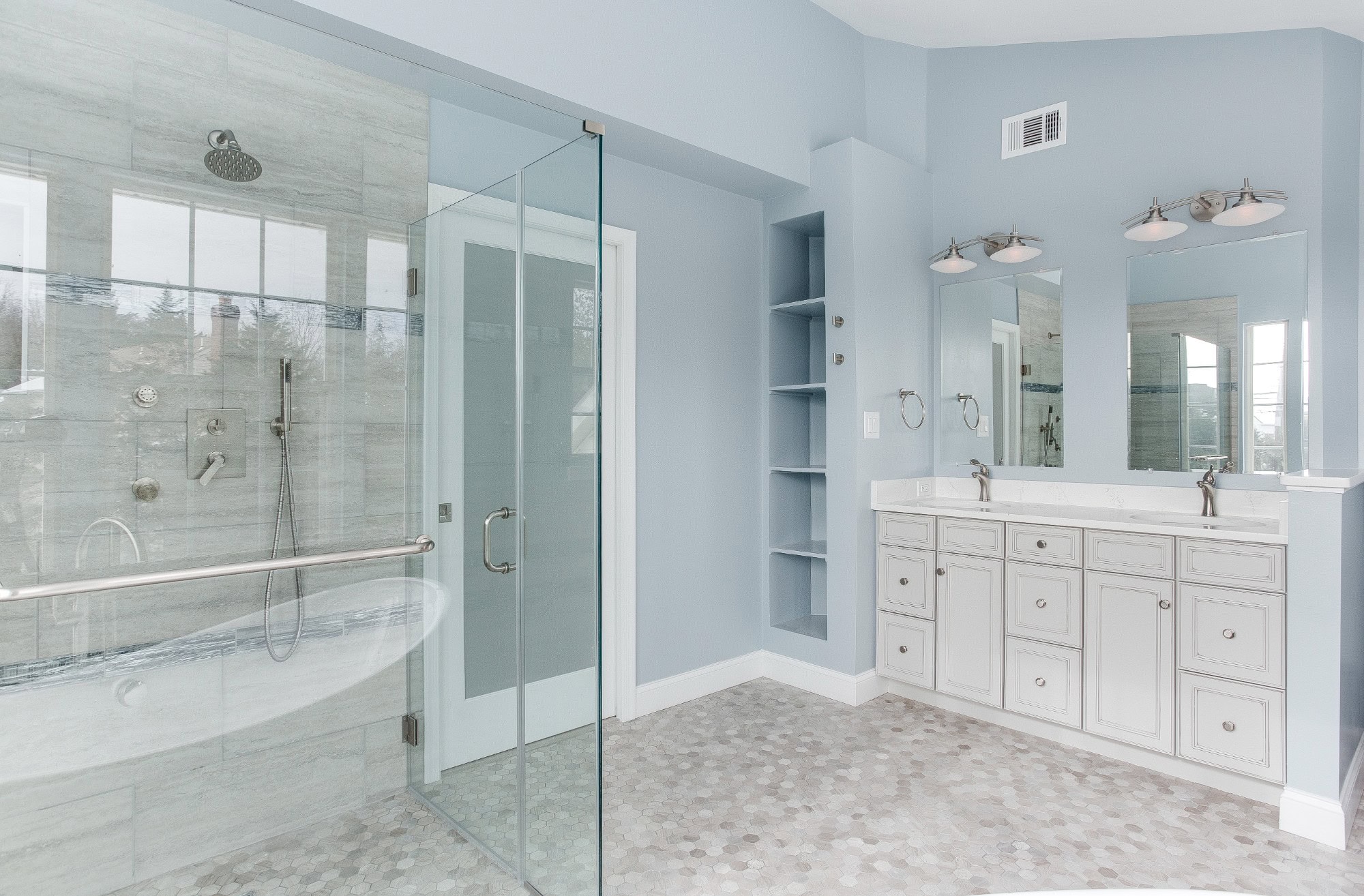 Light blue primary bathroom in Northern Virginia by EA Home Design with a large walk-in shower behind frameless glass, a handheld shower on a slide bar, a horizontal bar inside the glass, built-in shelving and a double vanity