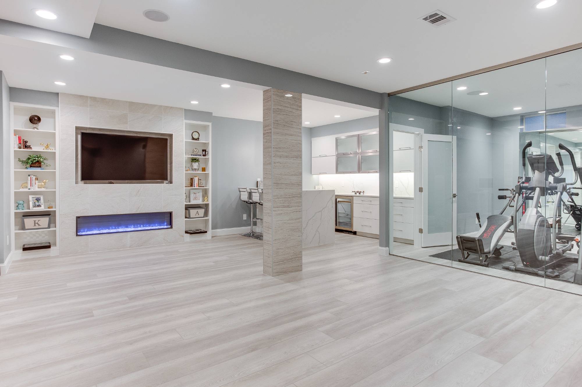 Finished basement in Northern Virginia with a media wall and fireplace, a wet bar, and a glass-walled home gym with cardio machines