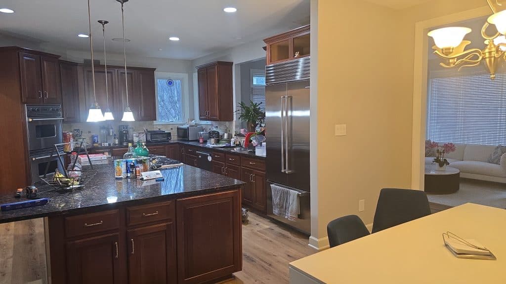 A modern kitchen with dark wood cabinets, granite countertops, and stainless steel appliances showcases stylish home improvement in Fairfax County, VA. The island has various items on it, with a dining table nearby and a living area visible in the background.