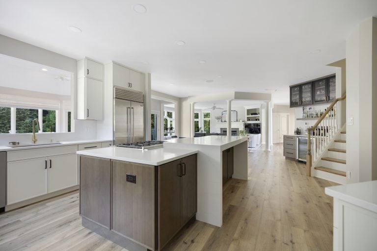 Modern kitchen with white cabinets, stainless steel appliances, a large island cooktop, and light wood flooring—perfect for your next home improvement in Fairfax County, VA. The space opens to a living area with stairs and a white railing visible on the right.