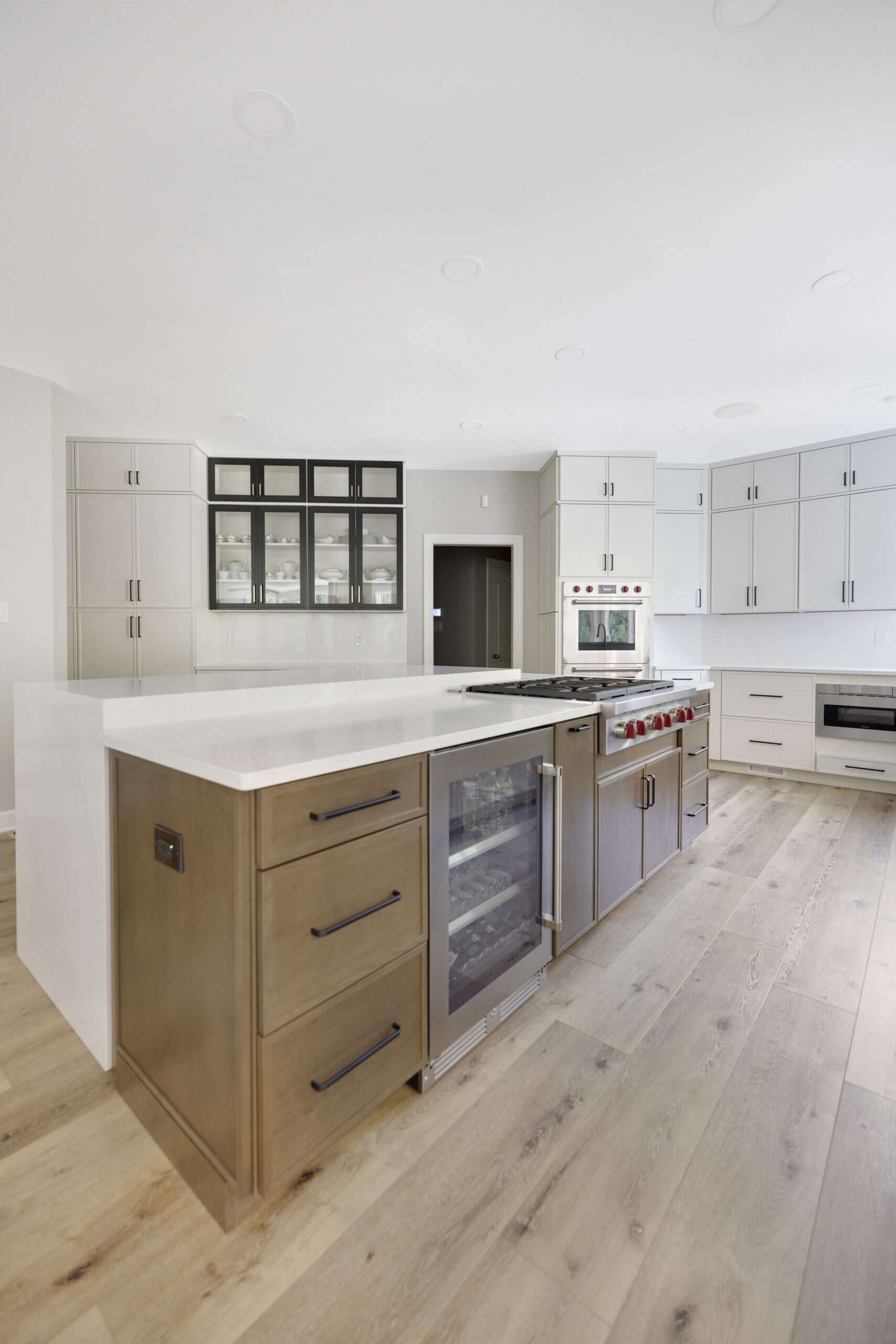 Modern kitchen with light wood floors, white cabinets, a spacious island, built-in stove, wine cooler, and stainless steel appliances—ideal for any home improvement in Fairfax County, VA. Glass-front cabinets and ample storage complete the look.