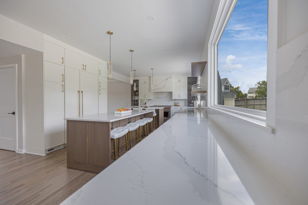 A modern kitchen features a long quartz island, bar stools, and a large window facing the backyard.