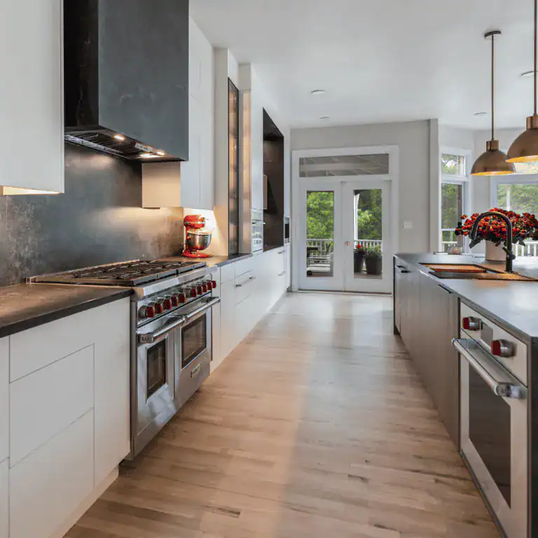 Modern kitchen with white cabinets, stainless steel appliances, a large island oven, pendant lights, and light wood flooring. Glass doors open to a patio with greenery—ideal for VA home improvement in Fairfax County.
