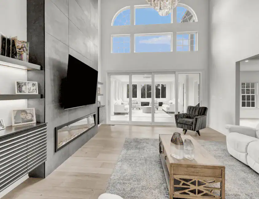 Modern living room with high ceilings and large windows, perfect for home improvement in Fairfax County, VA. Features a wall-mounted TV above a sleek fireplace, grey armchair, beige sofa, wooden coffee table, and shelves with decor and family photos.