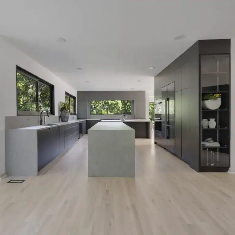 Modern kitchen with light wood flooring, large gray island, dark cabinets, built-in appliances, and big windows letting in natural light—an inspiring home improvement Fairfax County, VA homeowners will love. Decorative shelves showcase plants and dishes.