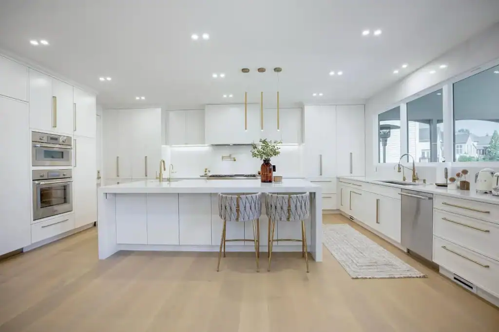 Modern kitchen with white cabinets, gold accents, a large island, built-in ovens, and wide windows for natural light. Light wood floors and minimalistic decor create a clean atmosphere—perfect inspiration for home improvement in Fairfax County, VA.