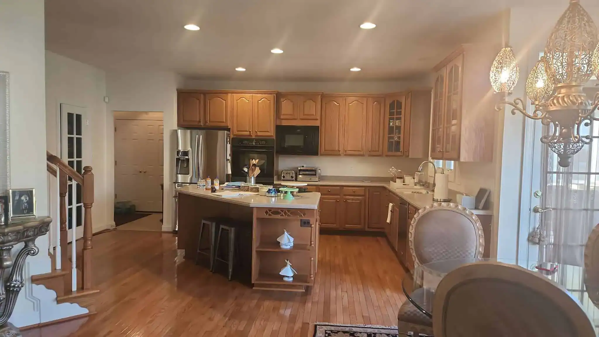 A spacious kitchen with wooden cabinets, stainless steel appliances, and an island countertop cluttered with various items—perfect for your next home improvement in Fairfax County, VA. Hardwood floors and ornate dining chairs complete the inviting space.
