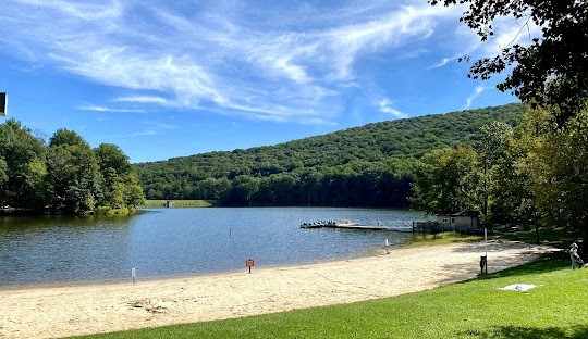A sandy beach leads to a calm lake bordered by trees, with a dock and small building on the right. Green hills rise in the background under a bright blue sky with wispy clouds.