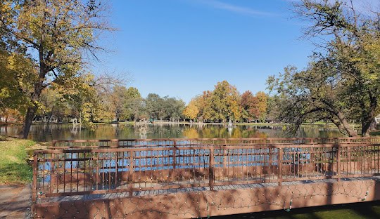 A wooden footbridge crosses over a calm pond surrounded by trees with green and autumn-colored leaves under a clear blue sky.
