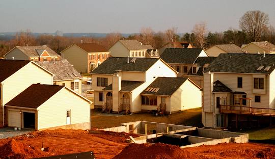 A suburban neighborhood with several two-story houses, some under construction. In the foreground, there is an exposed foundation and piles of red dirt. Trees without leaves are visible in the background.