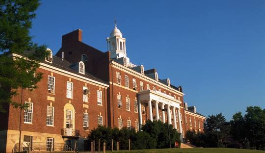 A large red-brick building with white columns and a cupola, featuring multiple windows and dormers, stands under a clear blue sky with trees and greenery in the foreground.