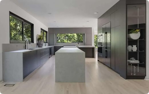 Modern kitchen with sleek, dark cabinets, light wood flooring, large center island, and big windows letting in natural light; a glass display cabinet with bowls and plants is on the right.