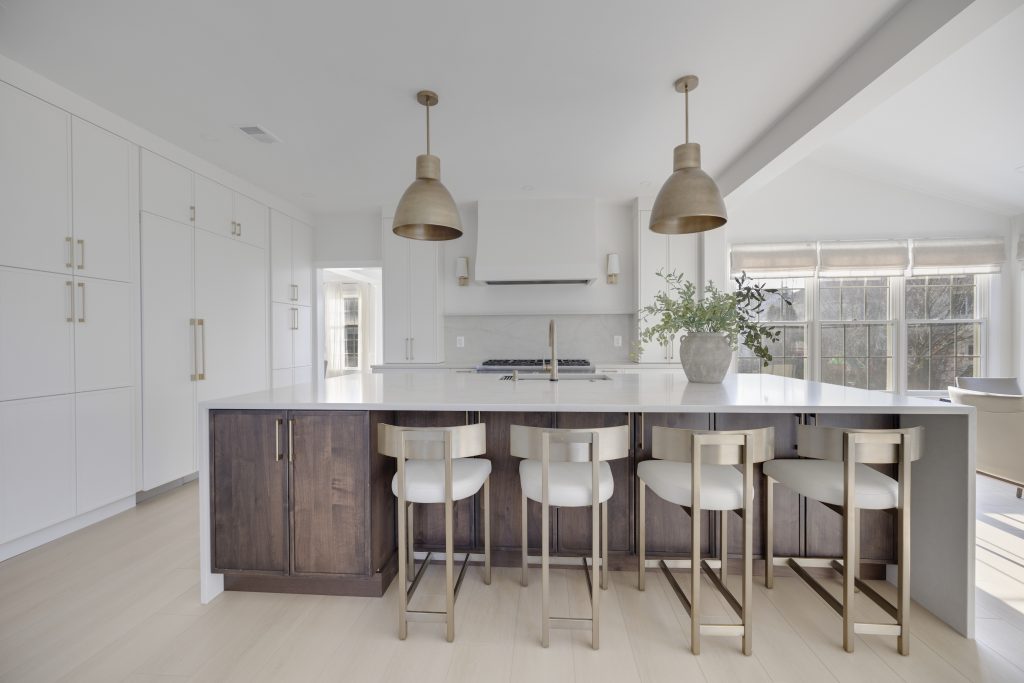 Modern kitchen with white cabinets, a large island with dark wood base, four white and gold bar stools, two pendant lights, and a vase with greenery. Sunlight streams in through large windows in the background.