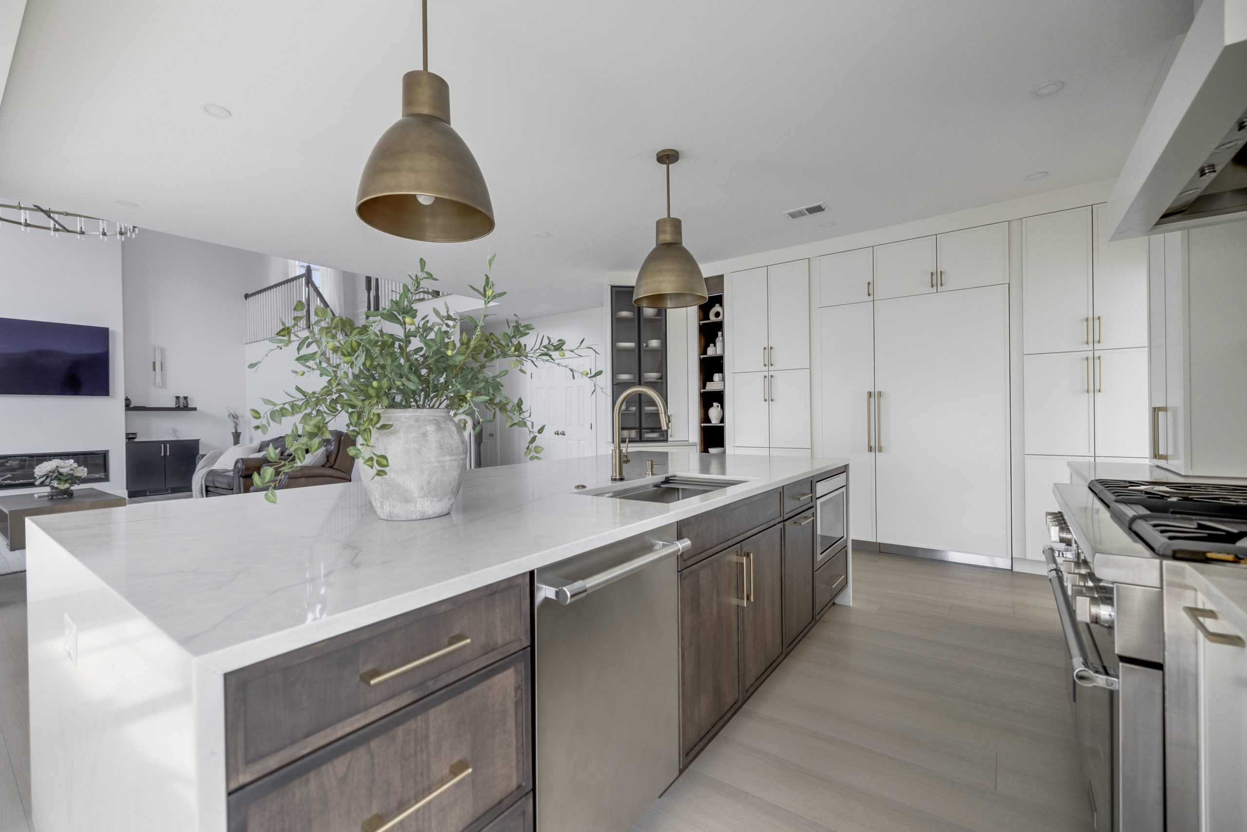 Modern kitchen with a large marble island featuring a built-in sink, dark wood cabinets, brass handles, and pendant lights. There is a potted plant on the island and white cabinetry in the background.