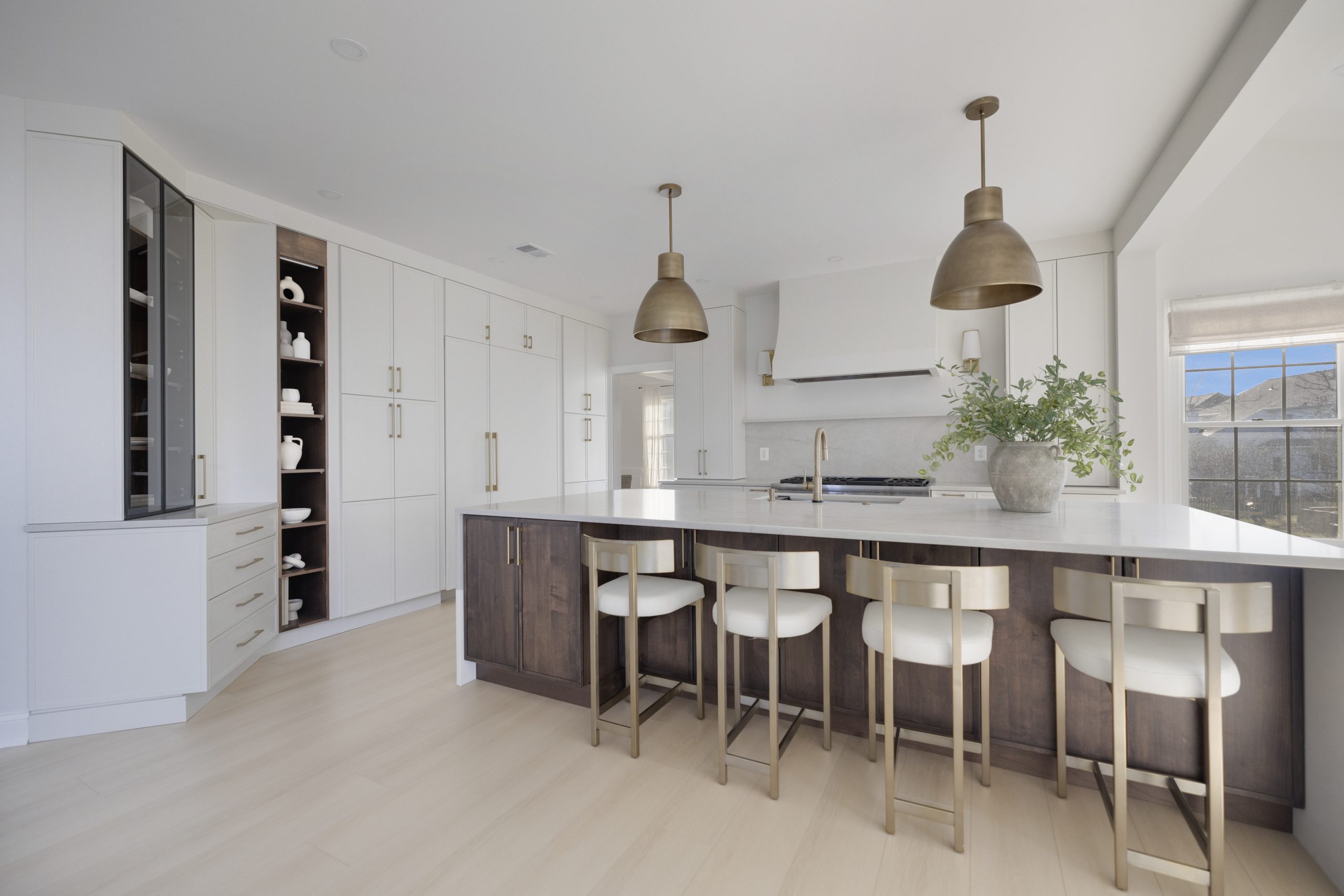 Modern kitchen with white cabinets, a dark wood island, gold bar stools, pendant lights, built-in shelves, and large windows letting in natural light. A potted plant sits on the island countertop.