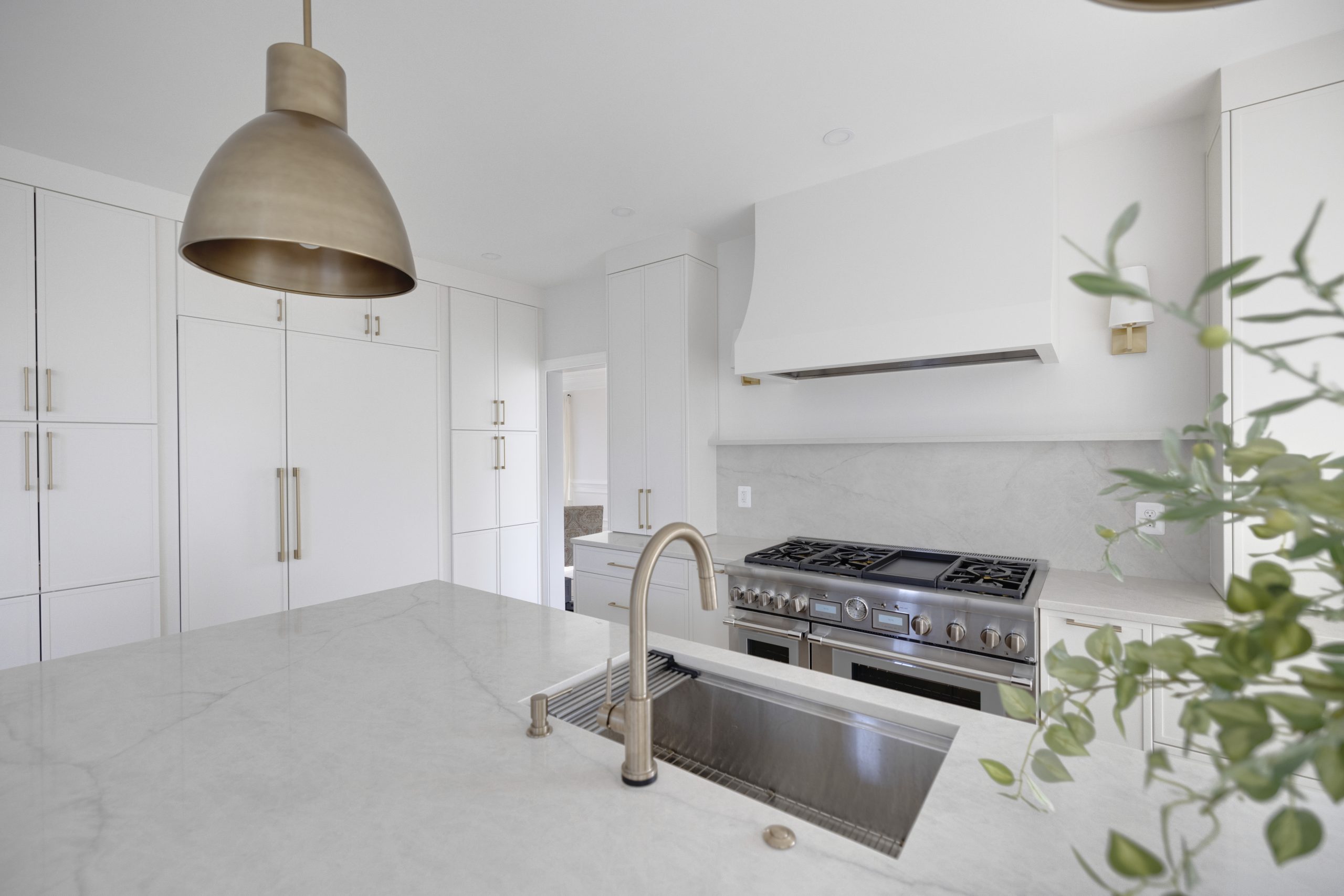 A modern kitchen with white cabinets, a marble countertop island with a built-in sink and gold faucet, a large gold pendant light, a stainless steel stove, and subtle greenery in the foreground.
