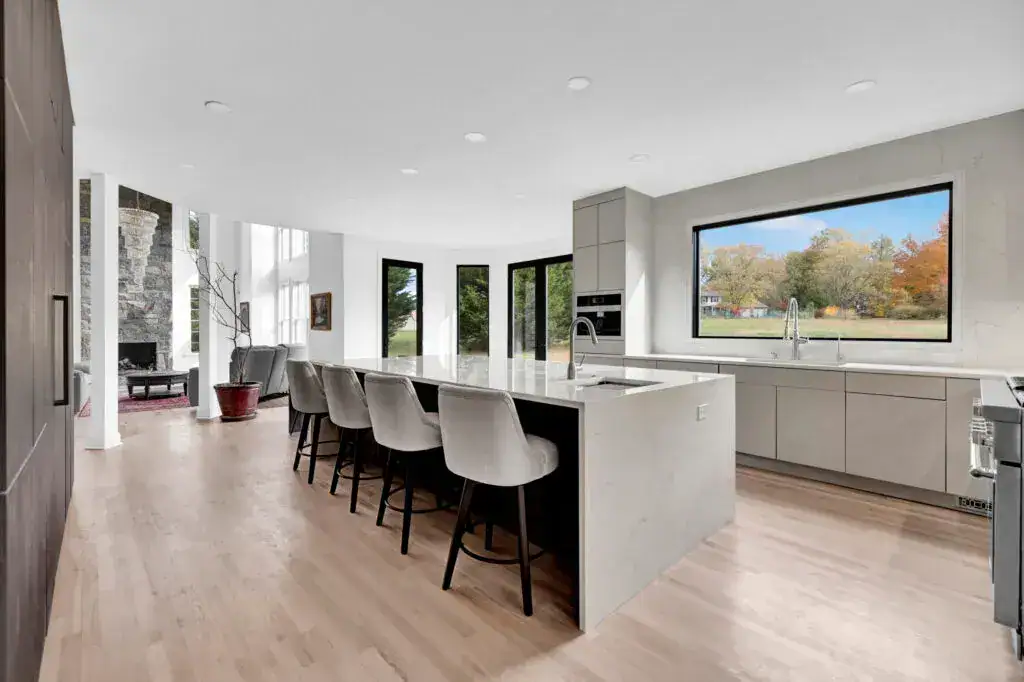 Modern kitchen with light wood floors, a large island with five upholstered chairs, minimalist light gray cabinets, a built-in oven, and a wide window offering a scenic view of trees and a lawn.
