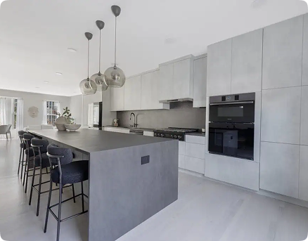 Modern kitchen with a large gray island, four black bar stools, minimalist cabinets, built-in oven, and three globe pendant lights. Bright, open layout with natural light from large windows.