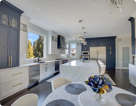 Modern kitchen with white and navy cabinets, a large marble island with barstools, stainless steel appliances, a sink under a large window, and a round dining table with placemats and a decorative centerpiece.