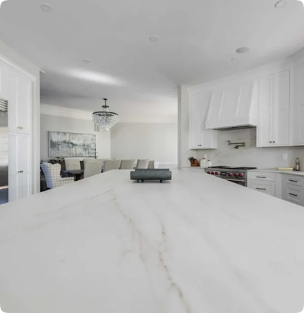 Modern kitchen with a large white marble island countertop, white cabinets, stainless steel appliances, and an open view to a dining area with a chandelier and abstract wall art.