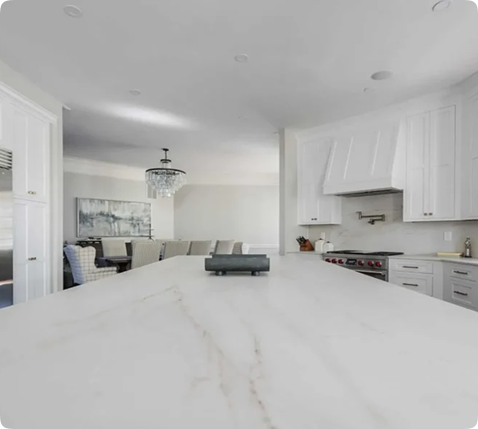 A modern kitchen with a large marble island countertop, white cabinets, stainless steel appliances, and a dining area with a chandelier visible in the background.
