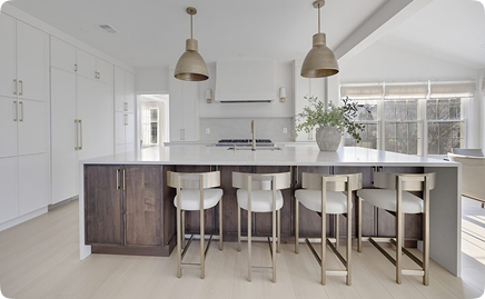 Modern kitchen with a large island featuring dark wood, four upholstered bar stools, white countertops, gold pendant lights, and a vase with greenery. White cabinetry and large windows bring in natural light.