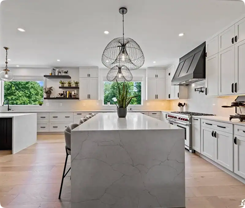 Modern kitchen with white cabinets, a large marble island with built-in seating, stainless steel appliances, pendant lights, a potted plant centerpiece, and large windows letting in natural light.