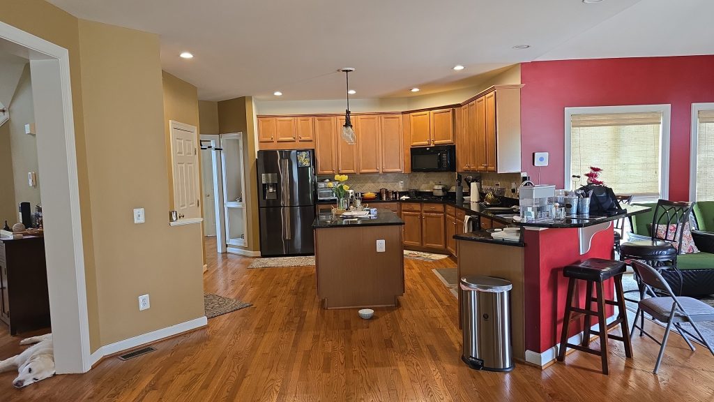 A spacious kitchen with wooden floors, light brown cabinets, and stainless steel appliances. The kitchen island has flowers and kitchenware. Bar stools and a trash can are near a red accent wall. A white dog lies in the hallway.