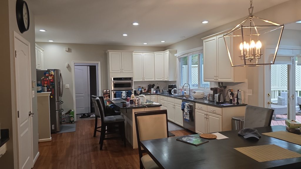 A spacious kitchen with white cabinets, stainless steel appliances, a center island with chairs, and various kitchen items on the counters. A dining table with chairs is in the foreground under a modern chandelier.