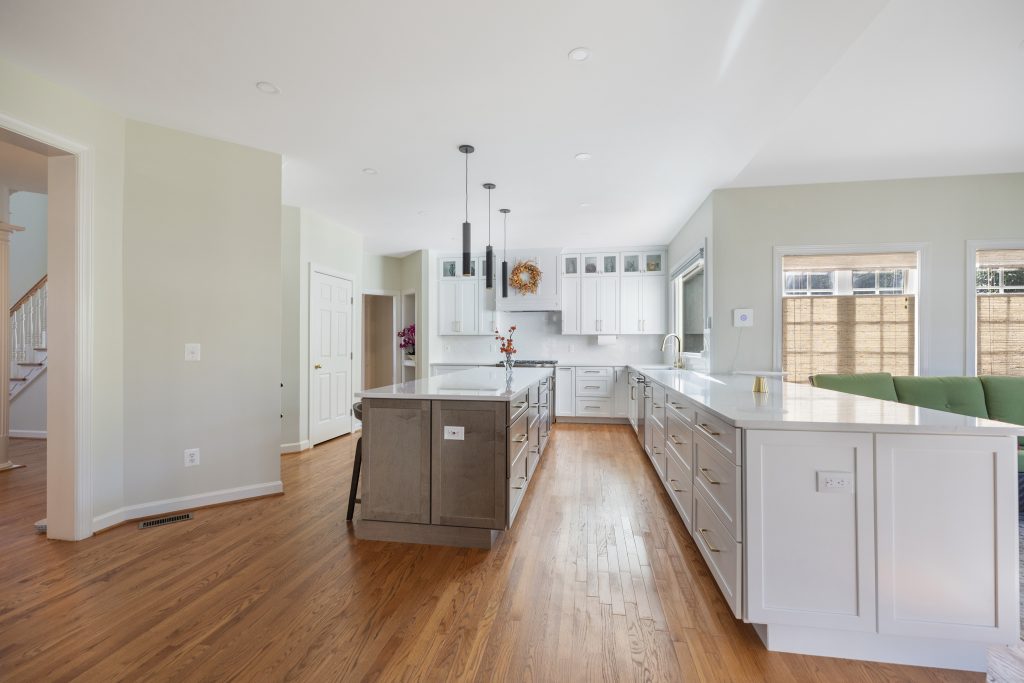 Spacious, modern kitchen with white cabinets, stainless steel appliances, a gray island with dark pendant lights, wood floors, and large windows letting in natural light.