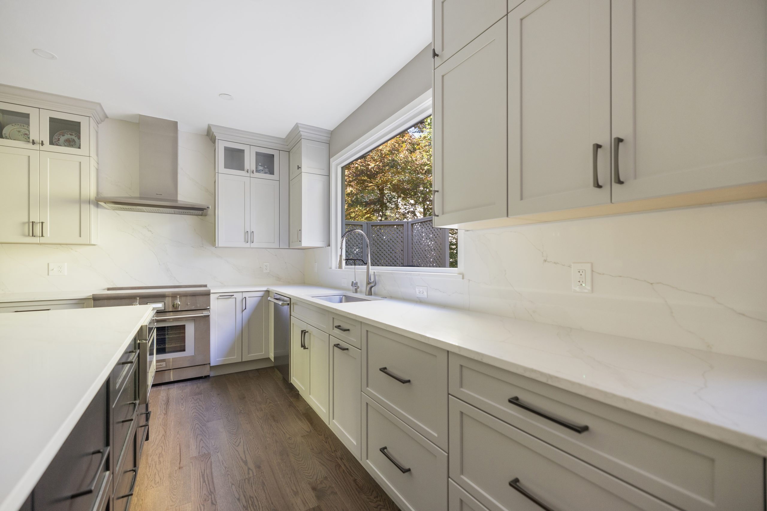 Light gray cabinets with white veined stone countertops and a matching full height slab backsplash behind the cooktop and under the window, in a Northern Virginia kitchen by EA Home Design