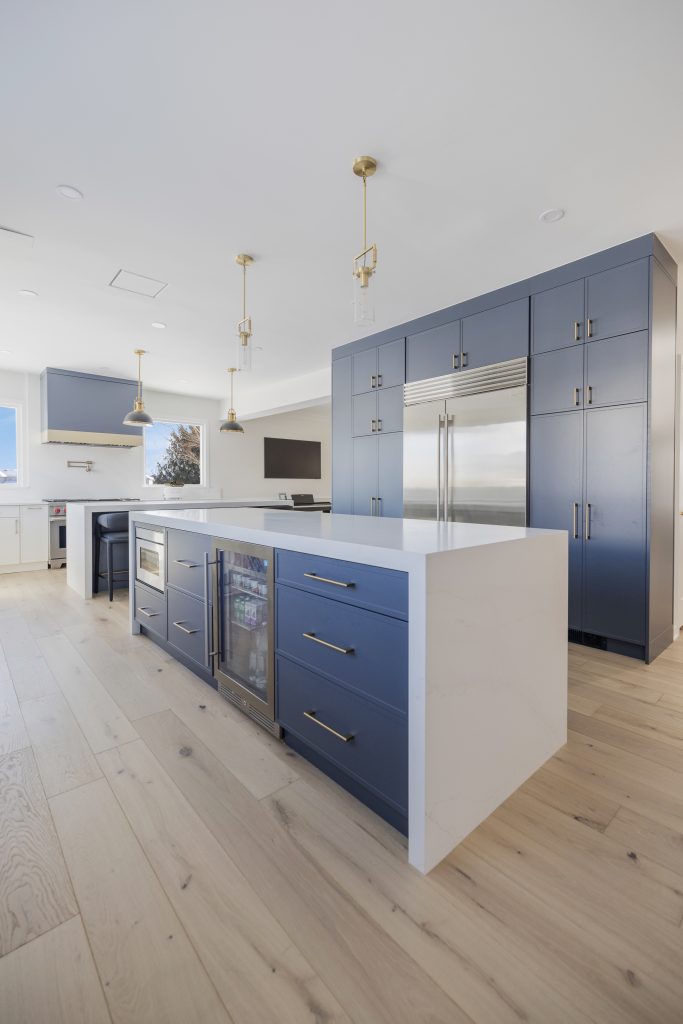 Modern kitchen with blue cabinets, a large white island, stainless steel appliances, and gold pendant lights. Light wood flooring and large windows create a bright, open space.